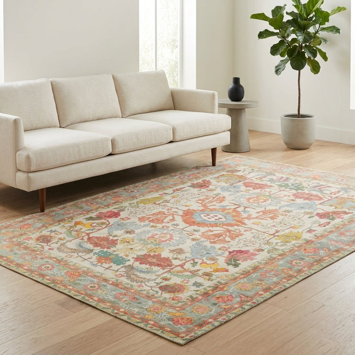 A rectangular area rug with a distressed traditional floral pattern in muted orange, blue, green, and beige tones, styled on a light wood floor in a bright living room next to a cream sofa and a potted plant.